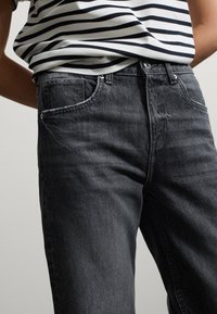 Jeans de mezclilla gris carbón con un ajuste relajado, cinco bolsillos y costuras visibles. Combinados con una camiseta de rayas en blanco y negro.