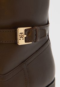 Brun læderstøvle med et spænde i guldfarve med præget logo, glat tekstur og struktureret design med syningsdetaljer.