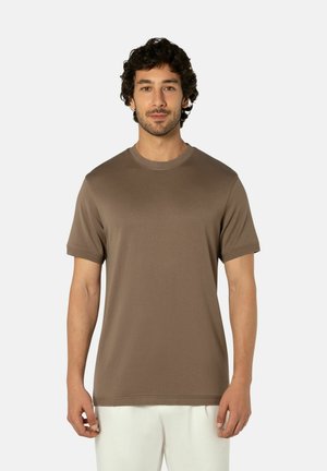 Man met kort krullend haar, gekleed in een eenvoudig bruin T-shirt met korte mouwen en witte broek, staand tegen een effen lichte achtergrond.