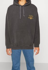 Felpa grigia scuro realizzata in morbido tessuto, con tasca a marsupio frontale, cordini e un logo ricamato in oro sul petto. Abbinata a jeans blu.