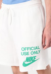 Valged puuvillased lühikesed püksid elastse vöökoha ja rohelise tekstiga "AINULT AMETIKS" ning Nike logo vasakul poolel.