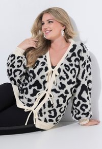 Mujer sonriente con cabello largo y rubio que lleva un cárdigan oversized de estampado de leopardo en blanco y negro con lazos crema, leggings negros y grandes pendientes de aro plateados.