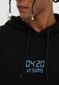 Black hoodie with digital print displaying "04:20 VI:SIONS" in light blue. Features drawstrings and metal aglets, textured fabric close-up.