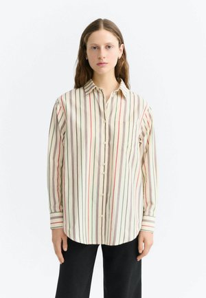 Femme aux longs cheveux bruns portant une chemise crème à rayures verticales rouges, vertes et marron et un pantalon noir, debout sur un fond blanc.