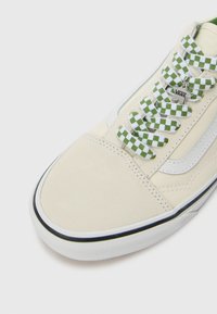 Beige canvas sneaker met groene en witte geruite veters, witte zijstrepen en een zwarte rubberen zool. Voorzien van een zilveren Vans logo tag.