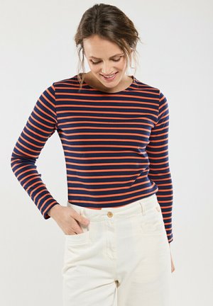 Chemise à rayures bleu marine et orange à manches longues avec un col rond, faite en tissu doux. Le modèle a la main dans une poche de pantalons blancs.