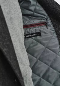 Premium wełniana kurtka o szarym wykończeniu, pikowana szara podszewka oraz bordowa kieszeń na zamek z etykietą "PREMIUM WOOL."