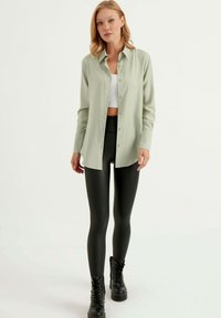 Camicia button-up di un verde chiaro con una vestibilità rilassata, indossata sopra un crop top bianco, abbinata a leggings neri e stivali neri con lacci.