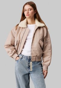 Veste bomber beige avec un col en shearling crème doux, fermeture éclair, poignets côtelés et étiquette. Portée sur un haut blanc et un jean bleu clair.