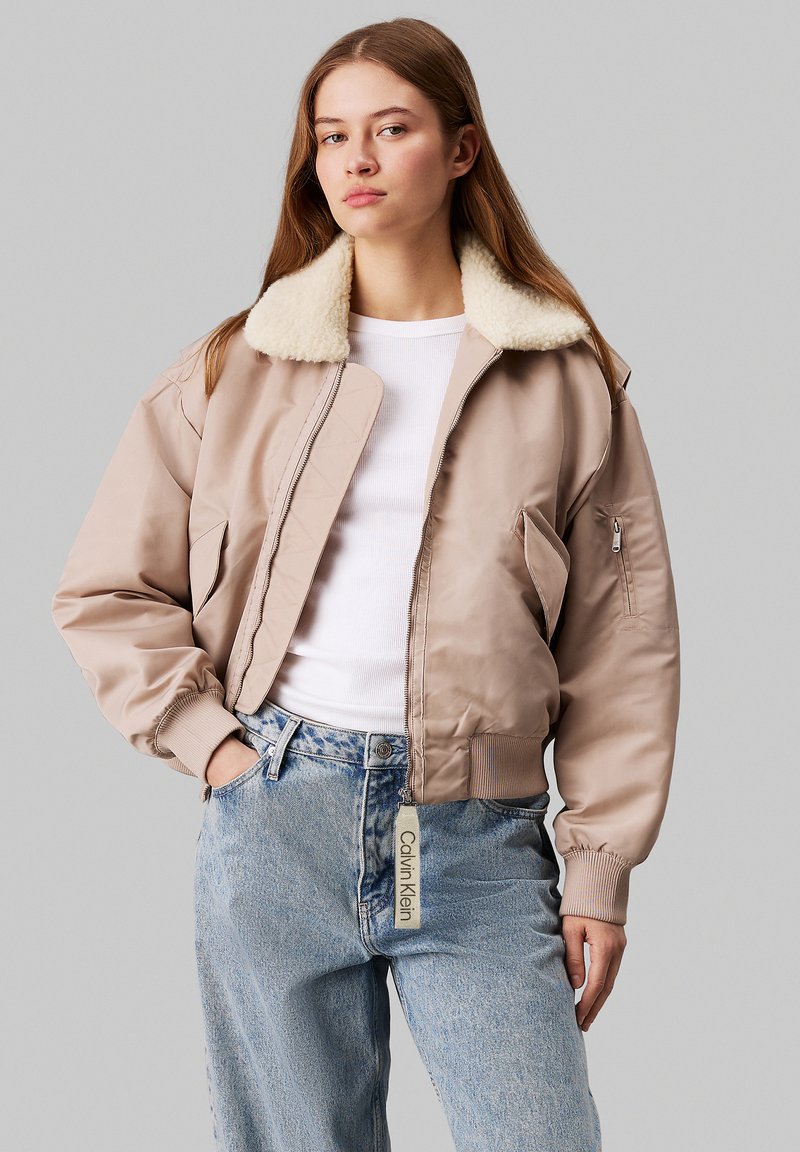 Veste bomber beige avec un col en shearling crème doux, fermeture éclair, poignets côtelés et étiquette. Portée sur un haut blanc et un jean bleu clair.