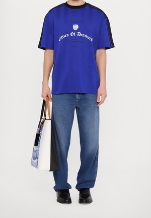 Persoon die een blauw "Citizen Of Denmark" T-shirt draagt, blauwe spijkerbroek, zwarte schoenen en een blauwe, witte en zwarte draagtas vasthoudt met bruine handvatten.