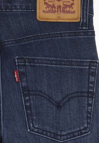 Jeans de mezclilla oscura con un bolsillo trasero visible, que presenta un patrón de curvatura cosido. Incluye una etiqueta de cuero marrón con detalles de tamaño y marca.