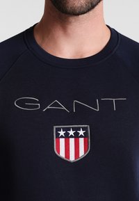 Felpa blu navy con scollo rotondo, caratterizzata da "GANT" ricamato in grigio, e un logo a scudo con strisce rosse e bianche e tre stelle bianche.