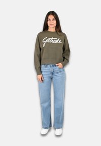 Pull cropped vert olive avec le texte blanc "Getrude", associé à un jean droit bleu clair et des baskets blanches. Tissu lisse, coupe décontractée.