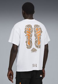 Hvid kortærmet t-shirt med et baggrundsdesign af brune og orange fodaftryk med stenlignende detaljer og logo.