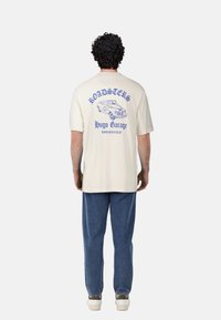 Cream-colored short-sleeve t-shirt featuring a blue graphic of a vintage car and text on the back, paired with blue denim pants.