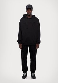 Sudadera negra con capucha y logo en relieve, combinada con joggers negros con estampado y zapatillas negras. El modelo se presenta contra un fondo claro.