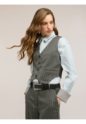 Vrouw met lang golvend haar, gekleed in een grijs krijtstreeppak met bijpassende broek, lichtblauw gestreept hemd en zwarte riem, kijkt opzij.