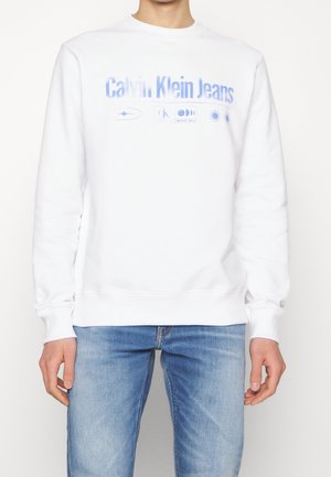 Osoba nosząca biały sweter Calvin Klein Jeans z niebieskim tekstem i symbolami, zestawiony z jasnoniebieskimi dżinsami.