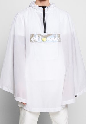 Personne portant un poncho blanc ample à capuche avec une demi-fermeture éclair et un patch logo réfléchissant à l'étiquette "ellesse" sur la poitrine.