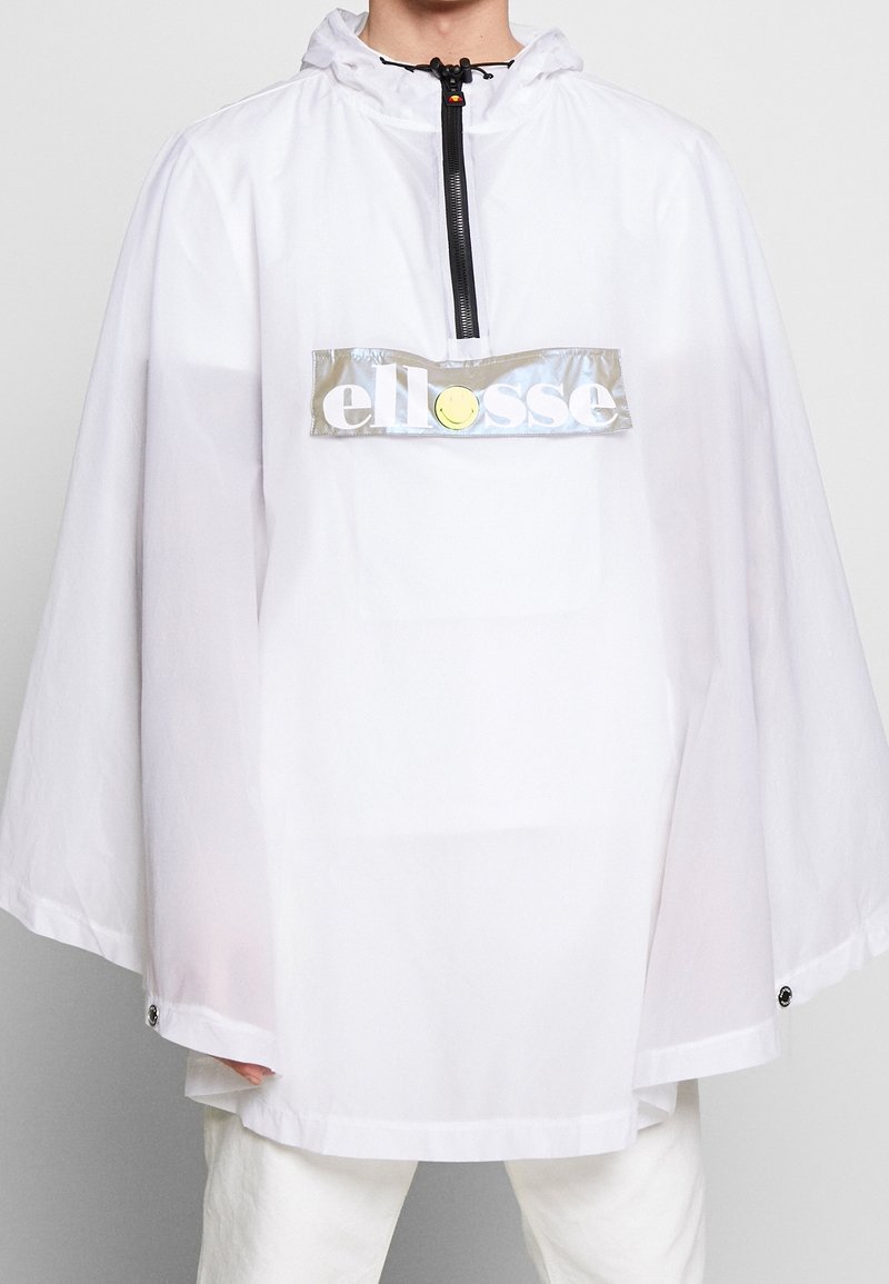 Personne portant un poncho blanc ample à capuche avec une demi-fermeture éclair et un patch logo réfléchissant à l'étiquette "ellesse" sur la poitrine.