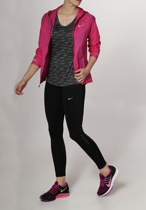 Veste légère rose à capuche sur un débardeur noir à motifs, leggings noirs et chaussures de sport multicolores avec des accents et des détails de logo.