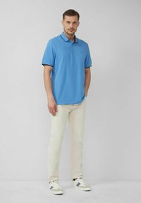 Helles blaues Poloshirt mit einem kontrastierenden dunklen Kragen und Emblem, kombiniert mit weißen Hosen und grauem Sneaker-Akzent. Glatte Baumwolltextur.