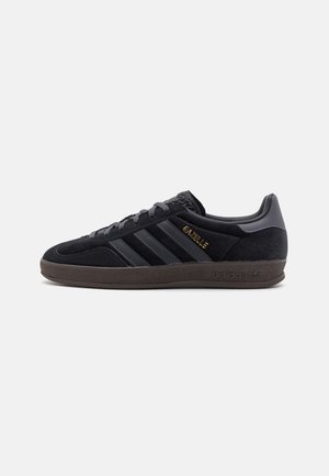 Czarny sneaker Adidas Gazelle z zamszową cholewką, trzema paskami, złotym logo oraz ciemnobrązową gumową podeszwą, widok z boku.