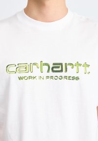 Witte katoen t-shirt met een groene graphic "carhartt. WORK IN PROGRESS" logo over de borst in een vet, modern lettertype.