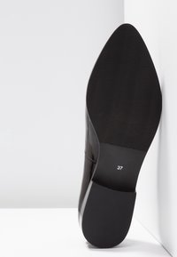 Semelle de chaussure élégante noire avec grip texturé et numéro de taille 37, positionnée debout contre des murs et un sol blancs.