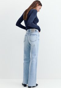 Femeie cu păr lung și șaten, purtând un top bleumarin cu mâneci lungi și blugi largi cu talie înaltă, de culoare albastru deschis, stând în picioare pe un fundal alb.