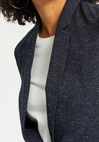 Marinblå blazer i texturerat tyg med en subtil glans, med snedskuren krage och öppen fram, bärs över en vit topp.