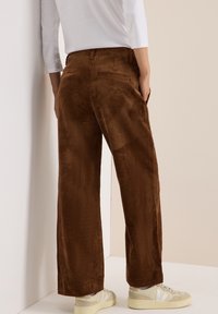 Bruine corduroy wijde broek met verticale ribbels, voorzien van twee achterzakken, gedragen met lichte sneakers.