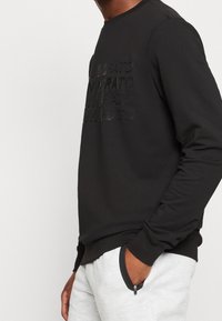 Sudadera negra con cuello redondo y manga larga, que presenta un texto en relieve en la parte frontal. La tela parece suave; el dobladillo y los puños son ajustados.