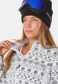 Biała polarowa kurtka z czarno-szarymi geometrycznymi wzorami, z kołnierzem na zamek. Czarna dziana czapka i gogle narciarskie w niebieskim odcieniu.