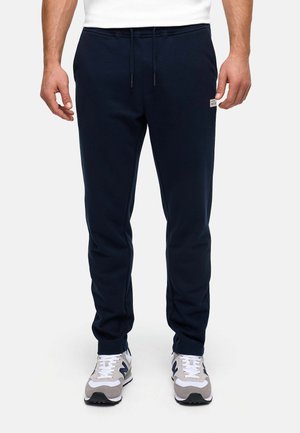 Uomo con pantaloni sportivi blu navy con coulisse in vita e scarpe da ginnastica bianche con dettagli grigi e neri, in piedi davanti a uno sfondo semplice.