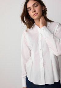 White button-up shirt made of cotton, featuring a classic collar, long sleeves, and a slightly pleated design with pearl buttons.
