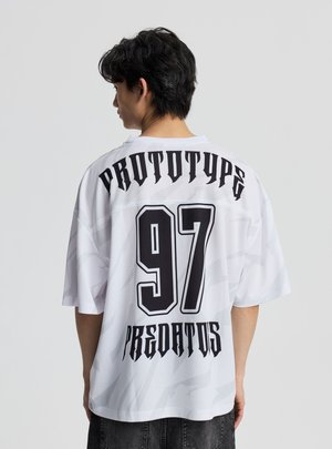Giovane con capelli neri che indossa una maglia bianca oversize con la scritta "PROTOTYPE," numero "97" e "PREDATORS" in carattere gotico nero.