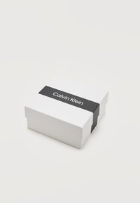 Λευκό κουτί με την επωνυμία Calvin Klein, με μαύρη ταινία που φέρει το λογότυπο της Calvin Klein στο κέντρο, σε απλό λευκό φόντο.