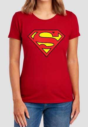Woman wearing red t-shirt with yellow and red Superman logo on the chest and blue jeans against a plain background.