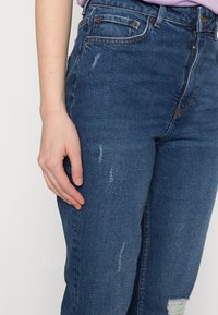 Calças de ganga em azul escuro com um acabamento desgastado, apresentando desgastes subtis e um design clássico de cinco bolsos. O material parece durável.