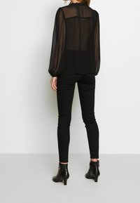 Musta, läpikuultava pusero pitkillä hihoilla, кerätty kaula-aukko ja pleatattu selkä, yhdistettynä tiukkoihin mustiin housuihin ja nilkkureihin.