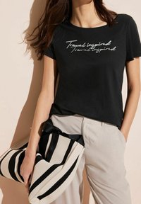 Černé tričko s krátkým rukávem, zdobené stříbrným nápisem "Cestování inspirováno." V kombinaci s světle béžovými kalhotami a pruhovanou taškou.