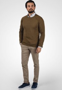 Brun genser med rund hals, brukt over en lys blå og hvit stripete skjorte, sammen med beige bukser og blå sko.