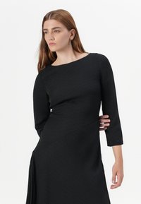A model wears a fitted black dress with three-quarter sleeves, displaying a confident expression against a white background.