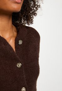 Braune, flauschig strukturierte Weste mit tiefem V-Ausschnitt und drei dekorativen goldenen Knöpfen auf der Vorderseite. Nahaufnahme vom Schulterbereich.