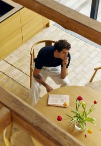 Jeune homme avec des tatouages assis pensivement sur une chaise en bois à une table ronde avec un journal et un vase de tulipes dans une pièce lumineuse.