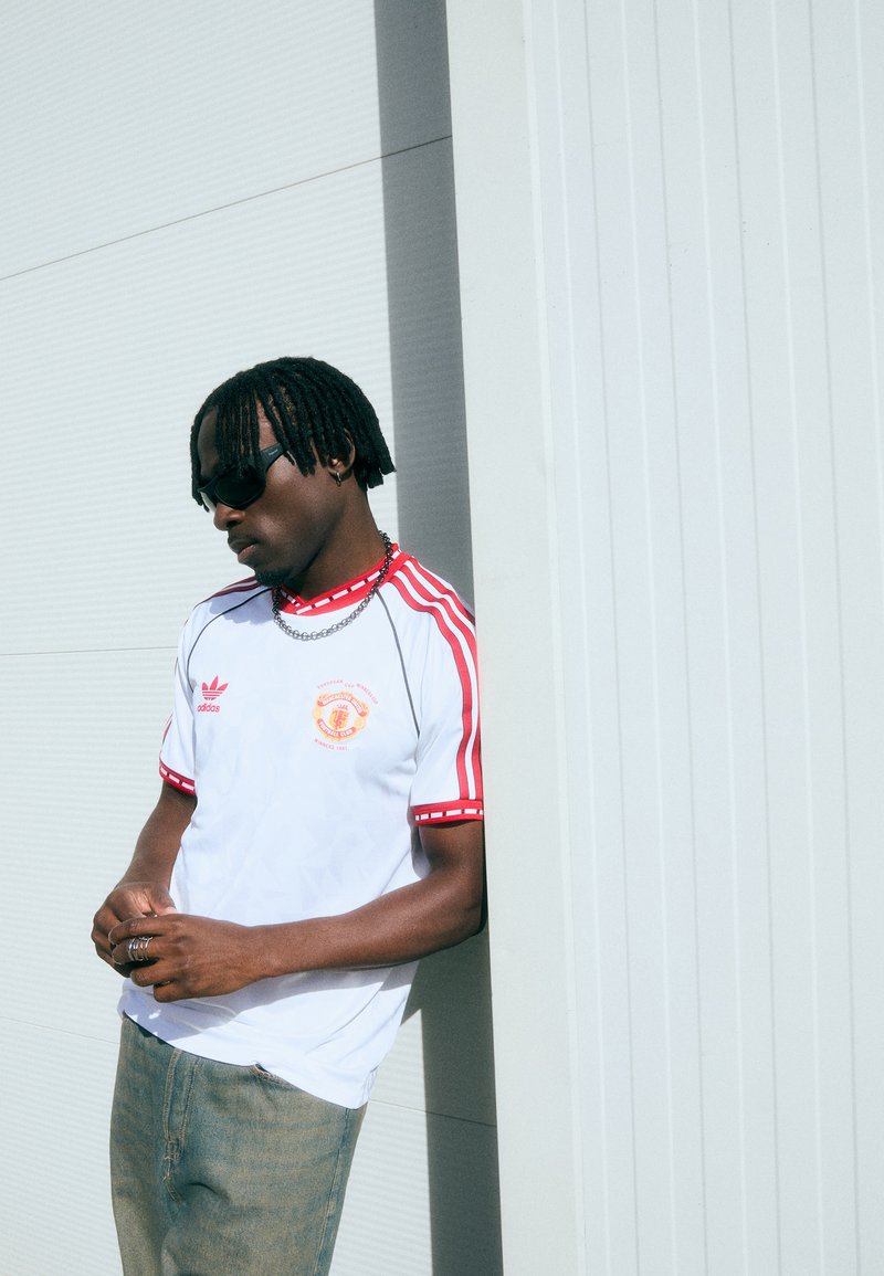 Vit idrottströja med röda detaljer och ränder, med en logotyp på bröstet. Strukturerad tyg och avslappnad passform kombinerat med ljusblåjeans.