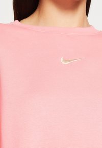 Sudadera rosa de Nike con cuello redondo y un peque ño logo dorado situado en la parte superior del pecho. Textura de tela suave.
