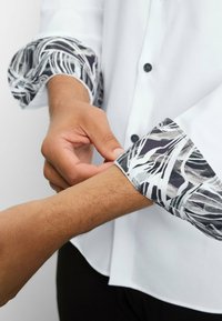 White button-up shirt with patterned black and gray cuff. Hand adjusting cuff, revealing cuff's textured design and detailing.